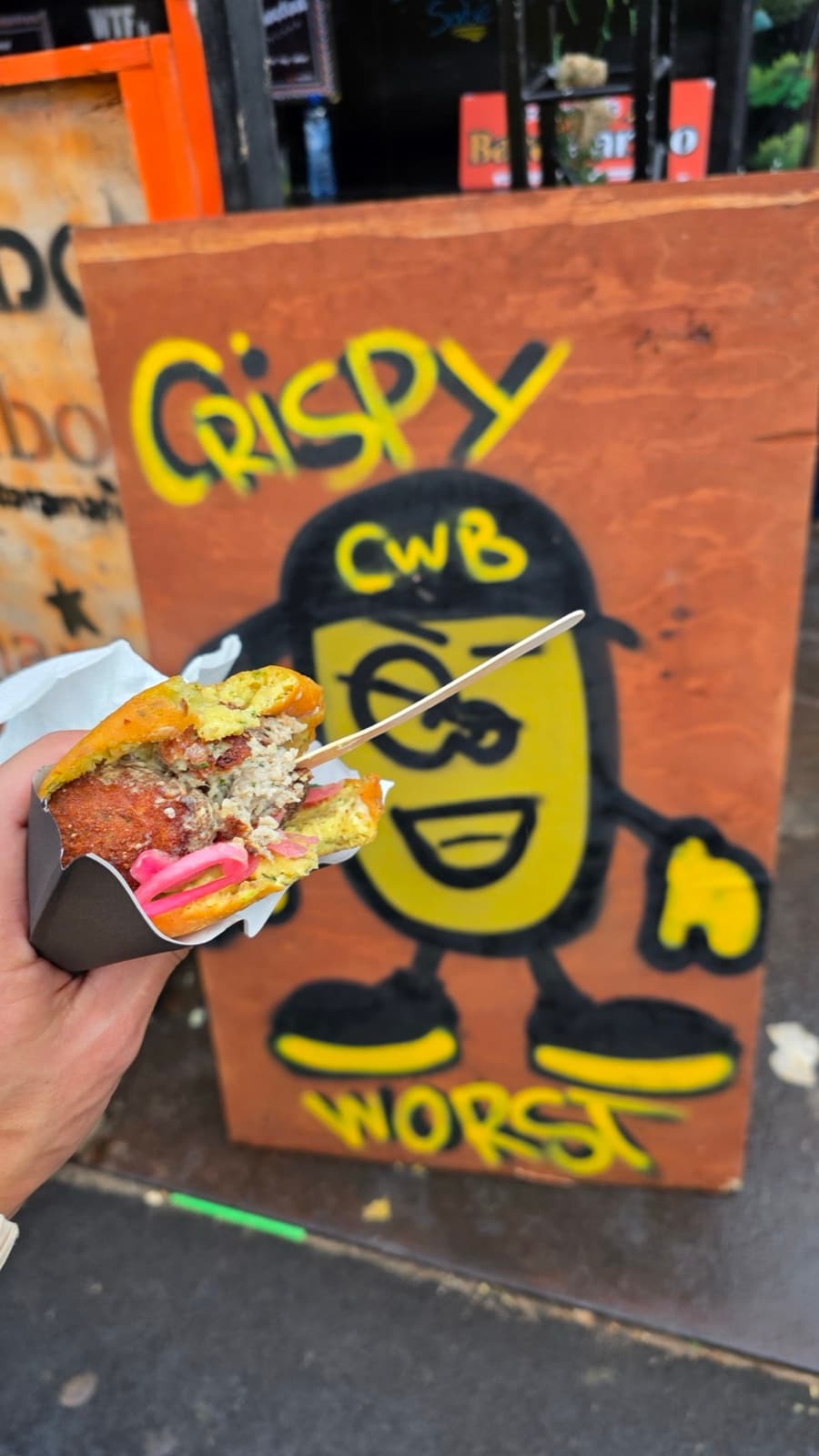 Broodje crispy vleesworst met roze zuur, vastgehouden voor een geschilderd Crispy Worst-bord op straat